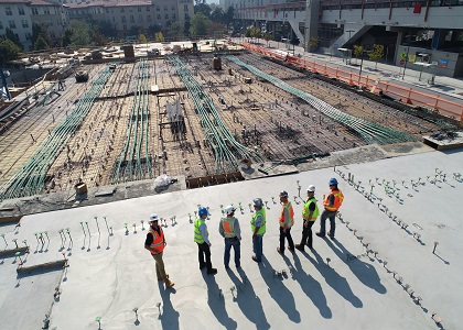 An aerial view of construction workers in hard hats and high-visibility vests standing on a large reinforced concrete foundation with exposed steel rebar.